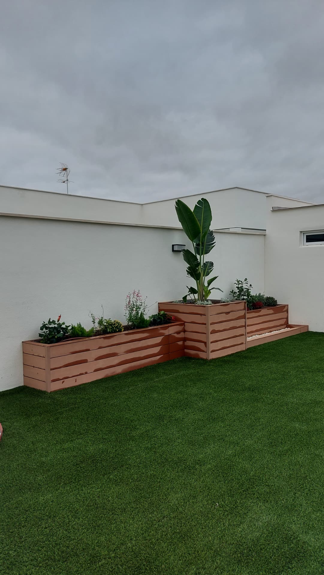 Jardineras de composite en L plantadas con strelitzia, hibiscos y aromáticas sobre césped